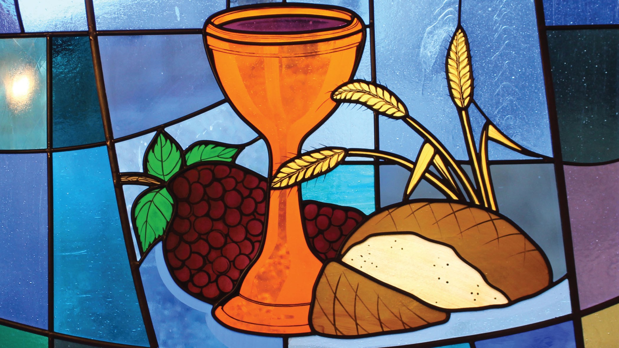 Stained glass of a copper-colored chalice filled with wine, a loaf of bread, a bunch of grapes, and stalks of wheat on a blue background