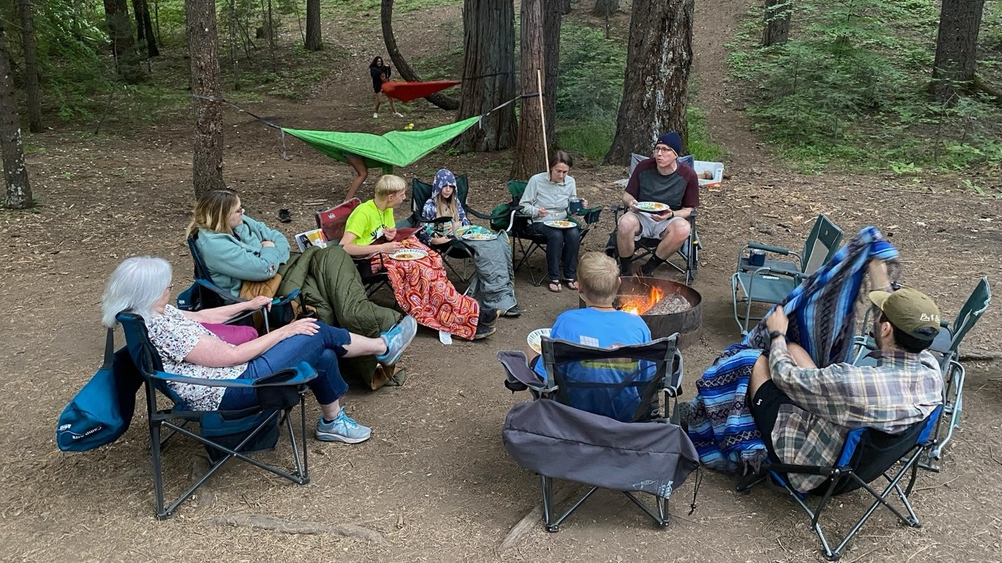 A group of people in folding chairs sitting around a campfire with pine trees in the background.